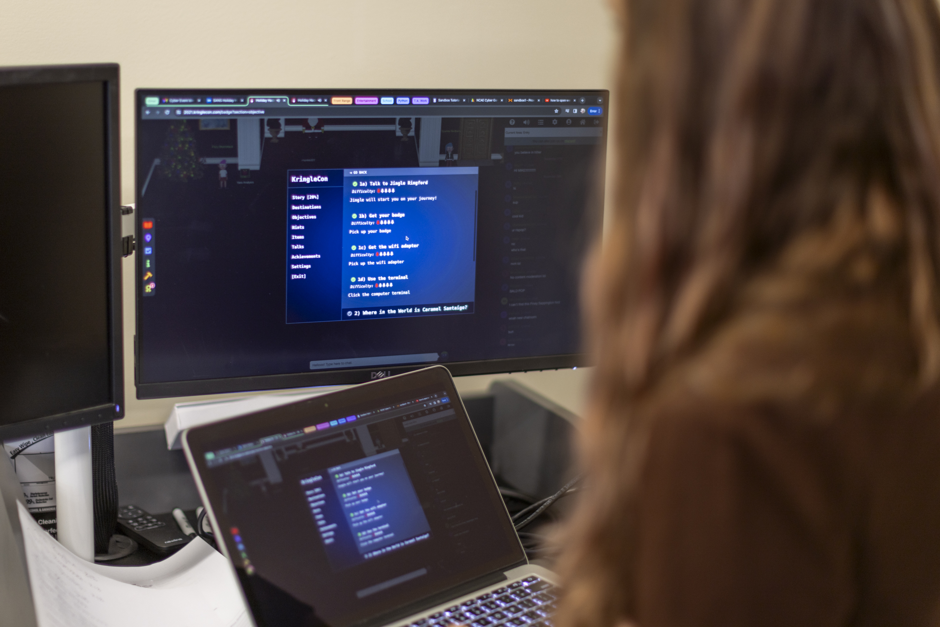 A picture taken over someones shoulder looking at a laptop screen and two desktop monitors. There is a computer program on the screens