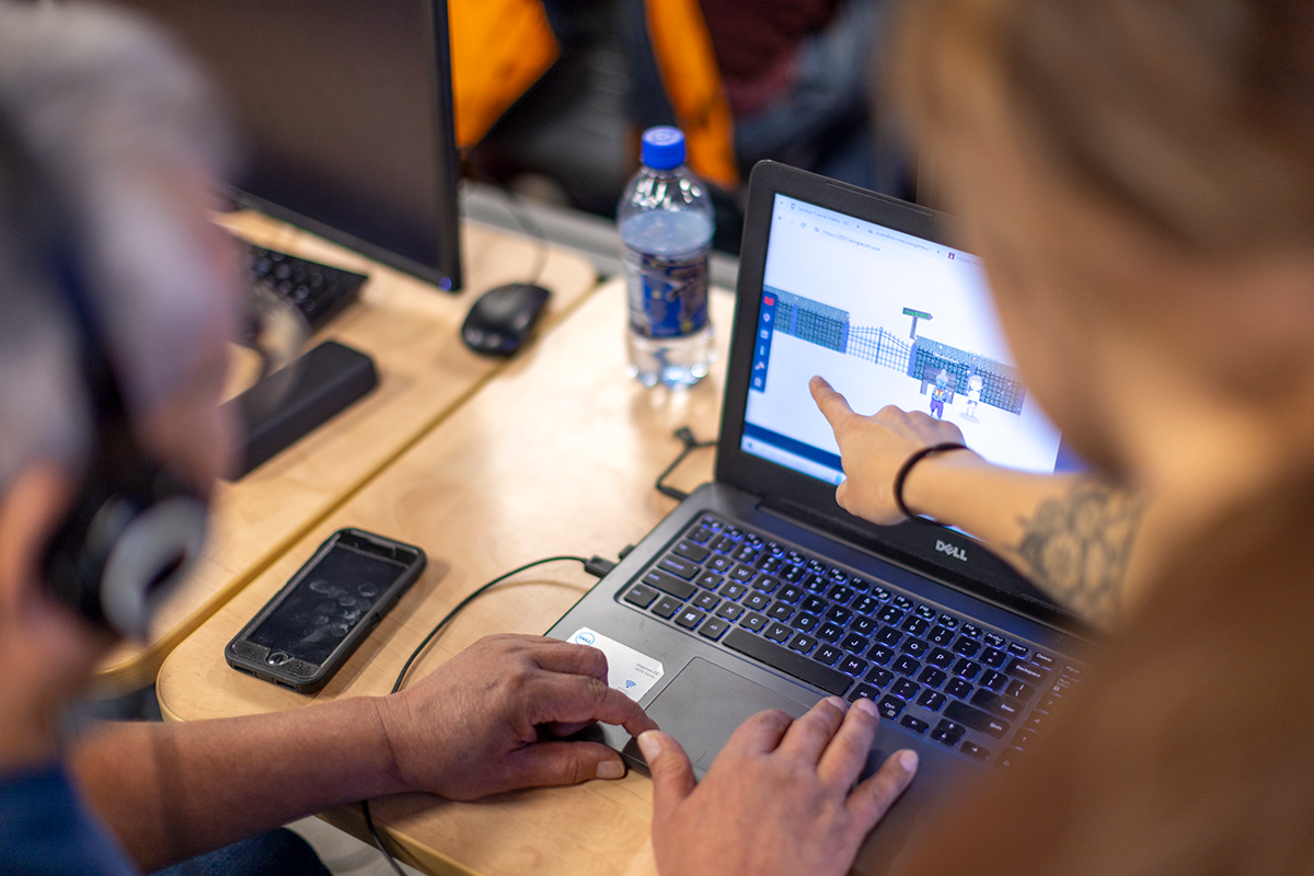 A person leaning over another person that's wearing headphones, pointing at something on a laptop screen