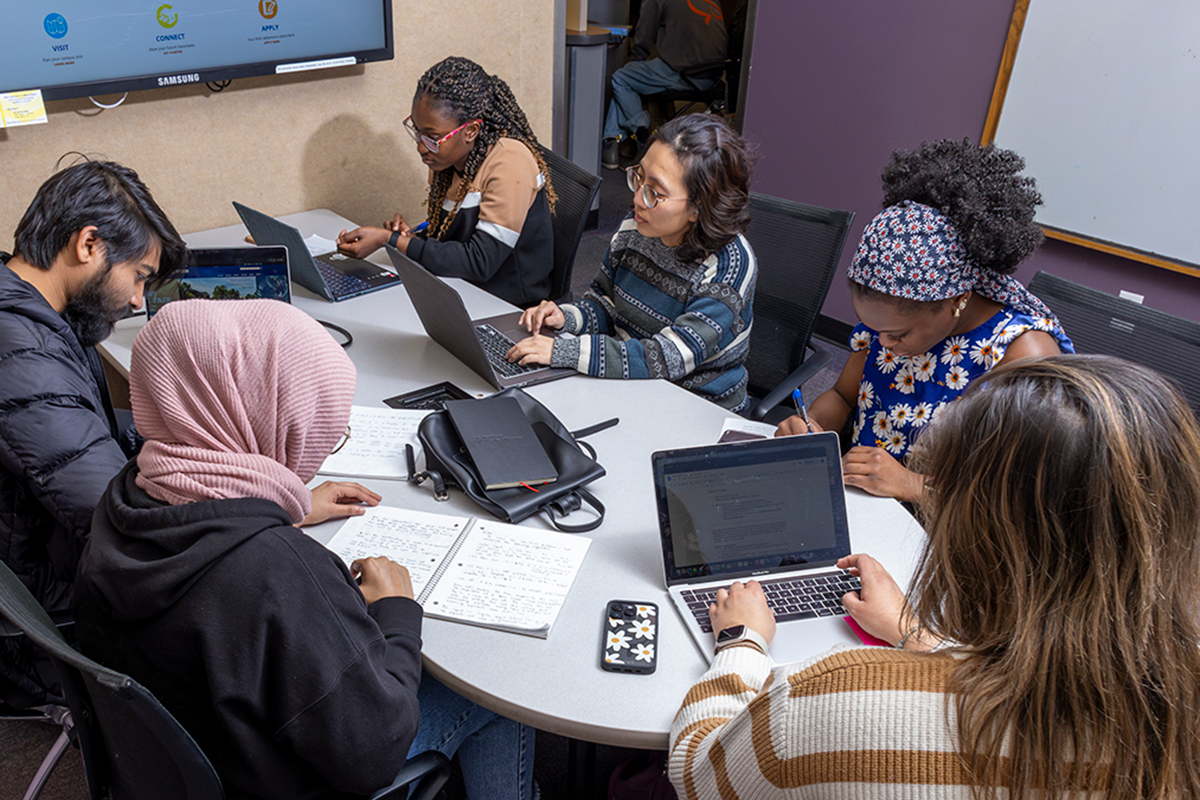 A group of students sitting at a table, typing on laptops and talking amongst each other