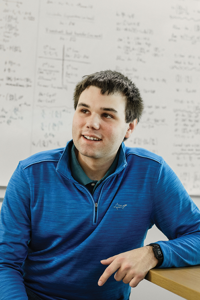 A person with short brown hair sitting at a table by a whiteboard