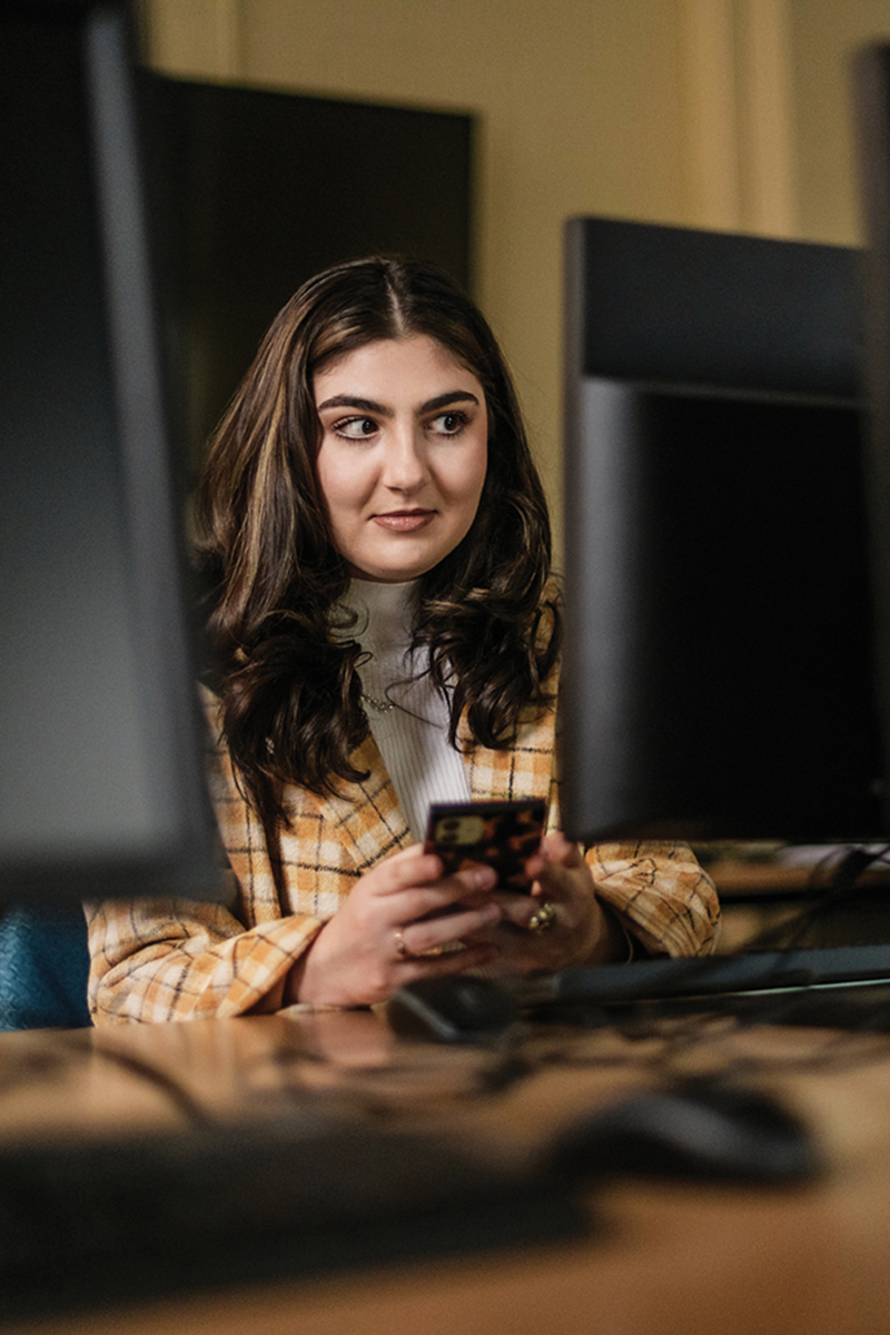 A person with long brown hair holding a rectangular device in a computer lab
