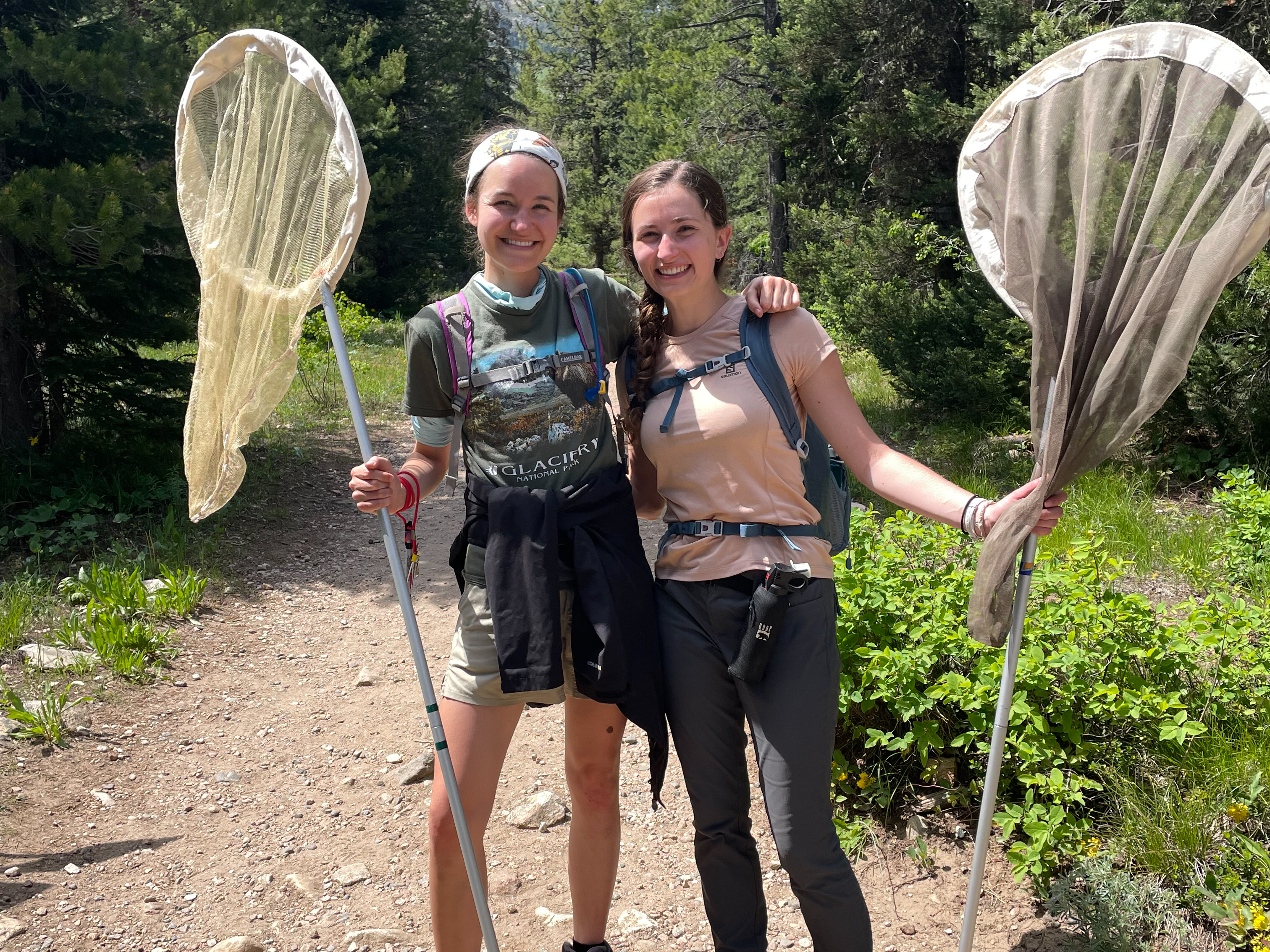 Two students posing arm-in-arm on mountain trail holding butterfly nets