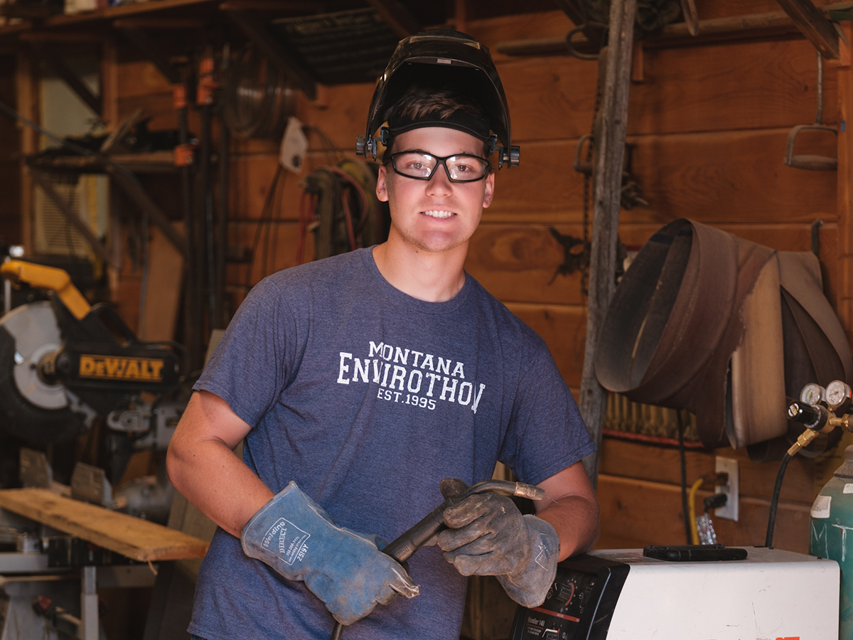 A person wearing a welding mask on the top of their head holding a blowtorch smiling to the camera