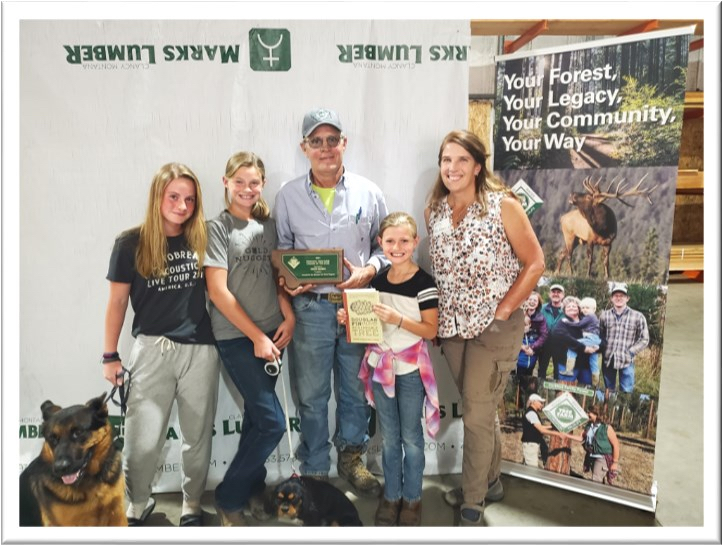 A group of five people and two dogs pose in front of a "Marks Lumber" backdrop, with the central man holding an award