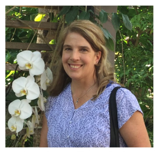 A smiling woman with shoulder length hair standing in front of a lush garden with orchids and green plants.