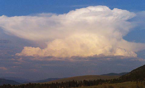 A large cumulonimbus thunderstorm cloud rises high above a landscape of rolling hills and scattered trees. The cloud has a towering vertical structure with a broad, flattened anvil-shaped top, characteristic of a mature thunderstorm.