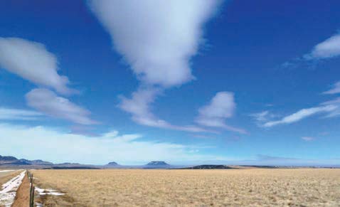 Elongated clouds stretch across a bright blue sky above a flat, open grassland. The smooth, layered cloud formations appear lens-shaped and aligned with atmospheric airflow over the landscape.
