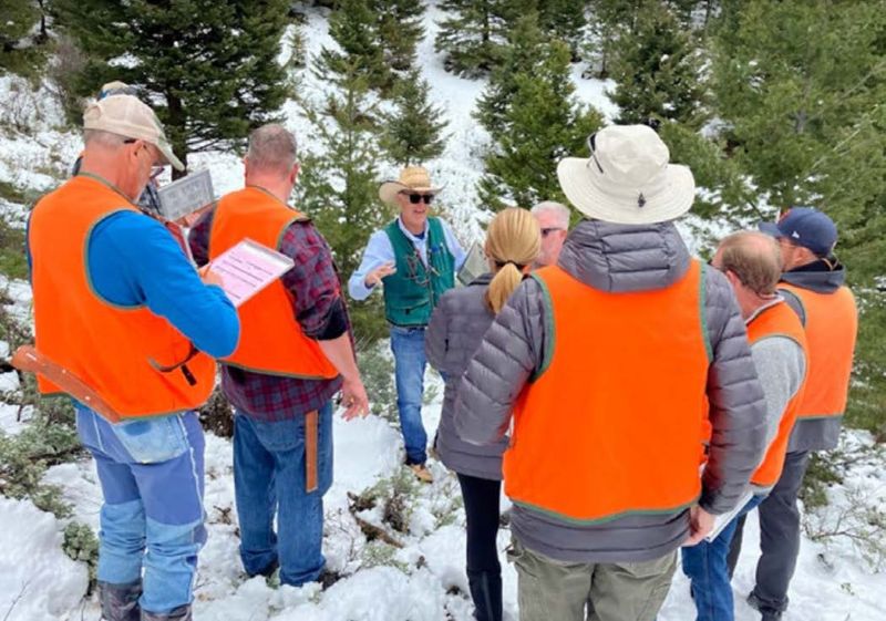 A group fo people in coats and orange vests stand in a forest and listen to a man in a cowboy hat and sunglasses.