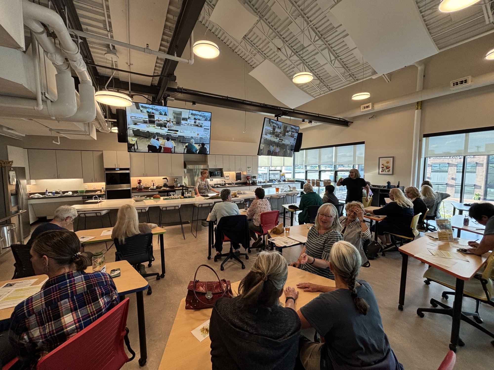 Participants at tables in a kitchen classroom watch a screen showing technique