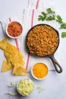 An image of lentil taco mix in a cast iron dish next to chips, onions, shredded cheese, and tomatoes.