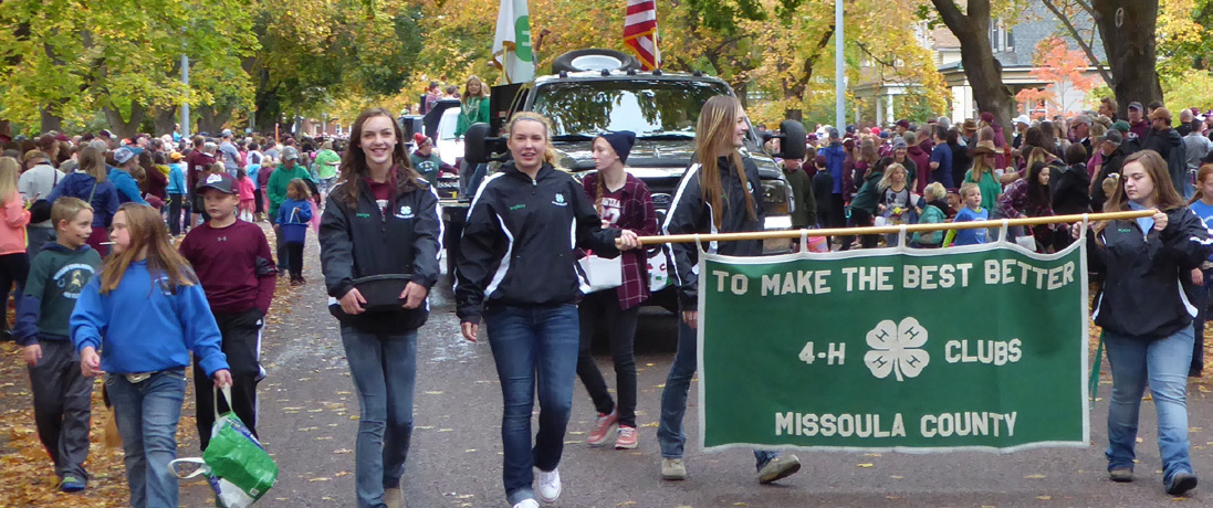 4-H members hold a 4-H banner while walking in the street during a parade. Crowds of people stand on both sides of the street.