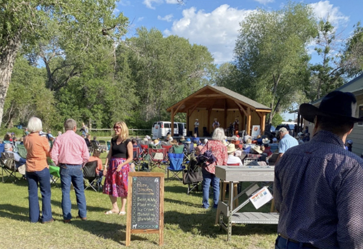 The community gathers on a nice summer day by a stage hosting a music group.