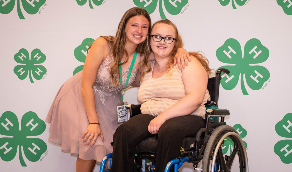 Two young women stand in front of a 4-H branded background. One of the ladies wears a pink dress and has her arm around the other who sits in a wheelchair.