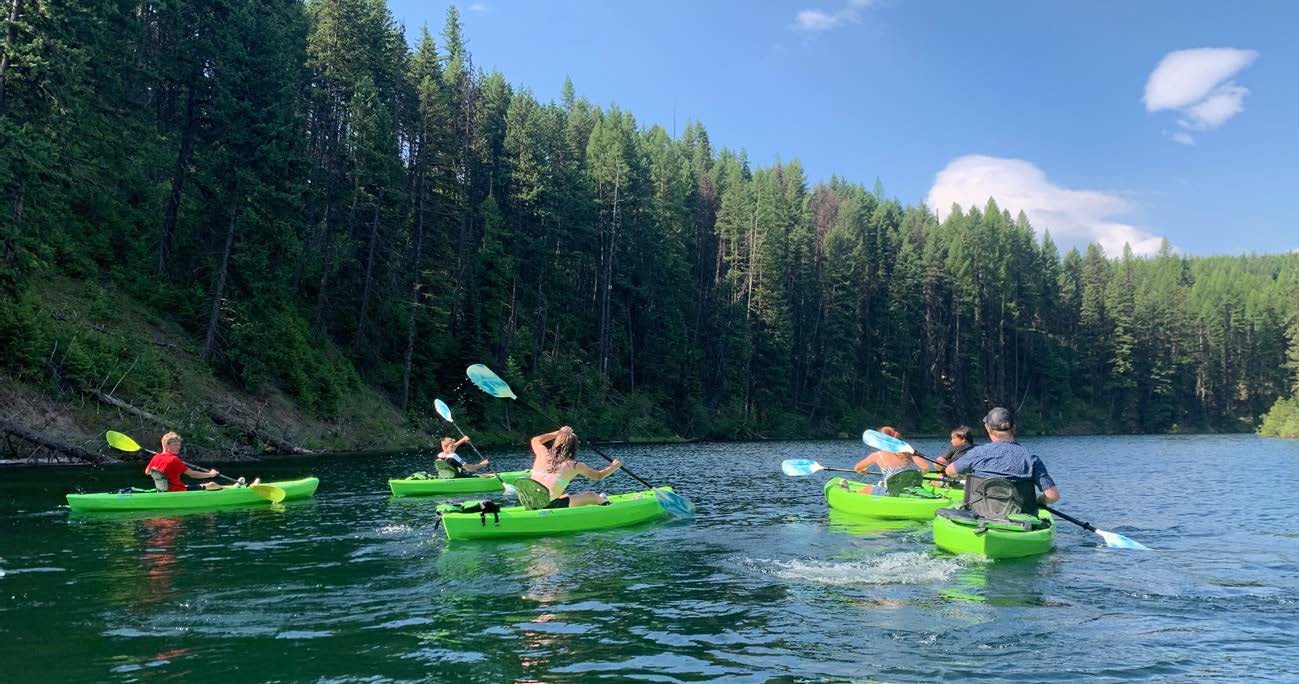 A group of young children kayak down a calm river surrounded by tall green trees, with an adult, all in green kayaks.