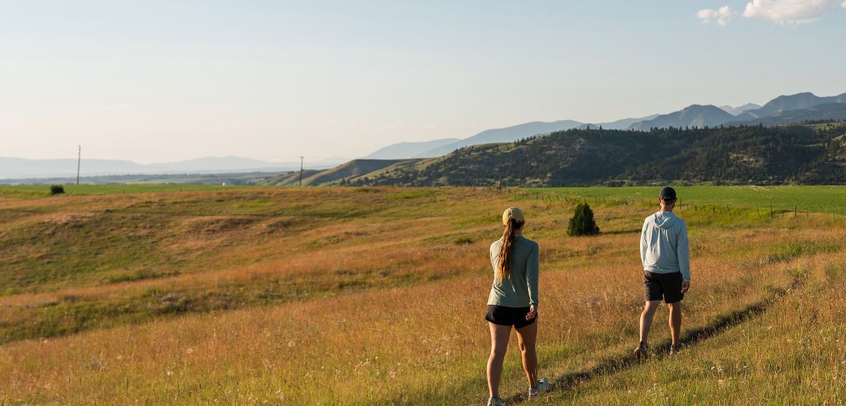 Two people walk on a trail through open grass. A fenceline and mountains are visible in the background.
