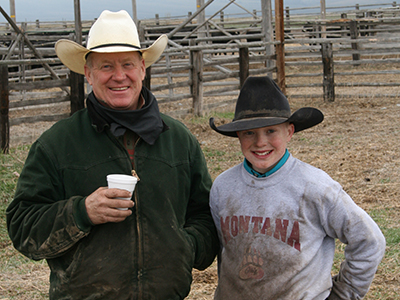 An older man and a boy stand side by side in front of a wooden corral and smile at the camera. Both are wearing cowboy hats and are covered in dirt.  