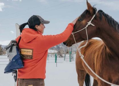 A woman holding skis and ski boots rubs the forehead of a horse wearing a lead.