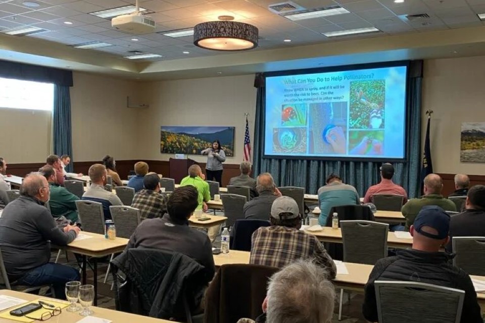 A group of people sitting and watching a speaker with a presentation about pollinators in the background.