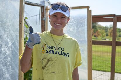 Volunteer wearing a Service Saturdays shirt and glove gives a thumbs-up while working outdoors near a greenhouse.