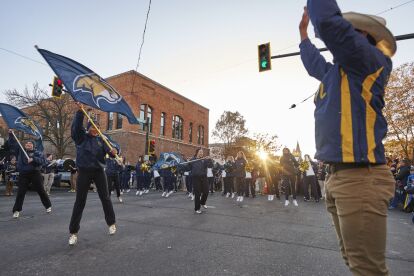 Marching band performs with flags and instruments on a city street during a homecoming pep rally at sunset.