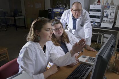 Two researchers in lab coats discuss data on a computer screen while a senior researcher observes.