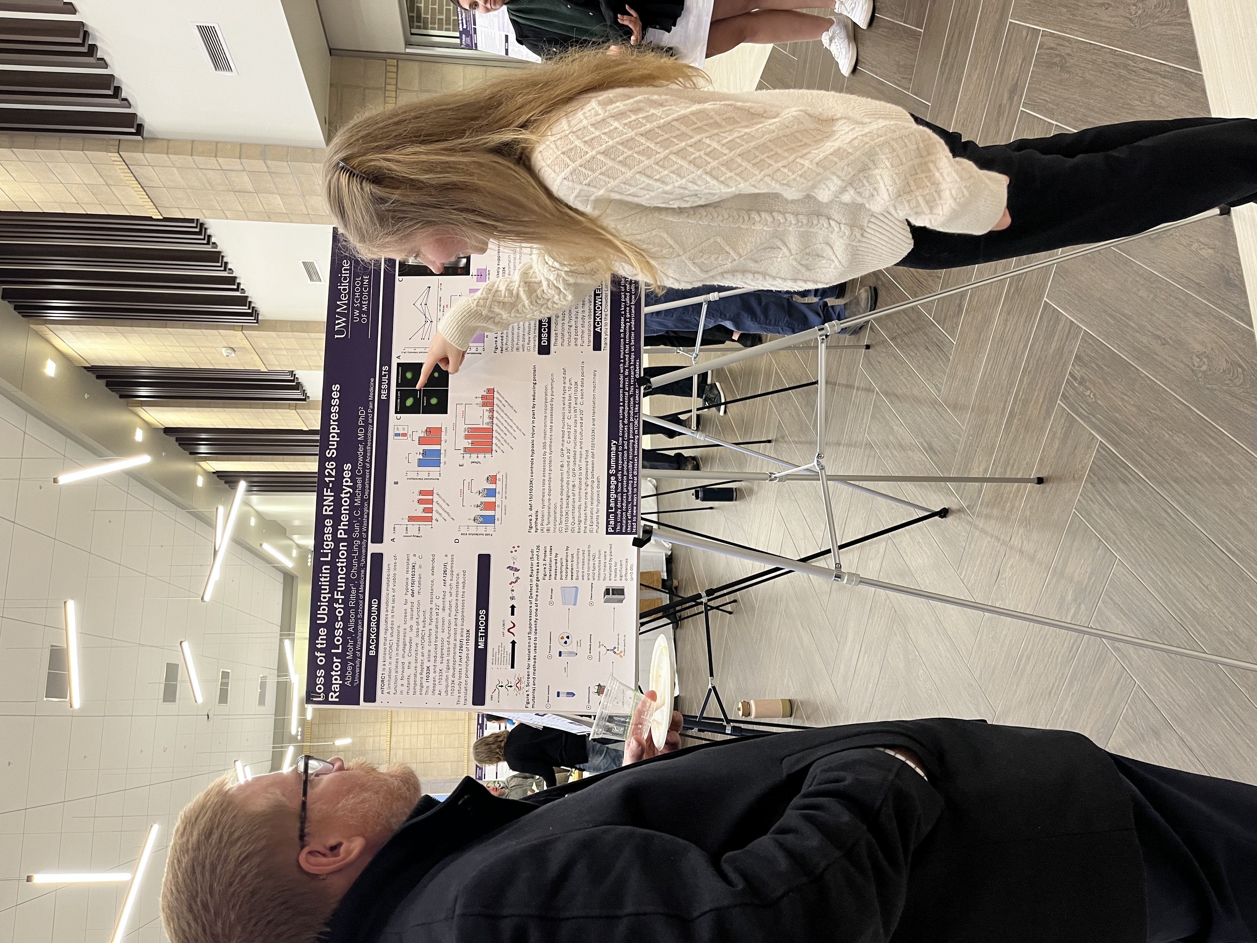 Poster Session 2025 Photo of a female medical student pointing to a chart on a research poster while a man looks on